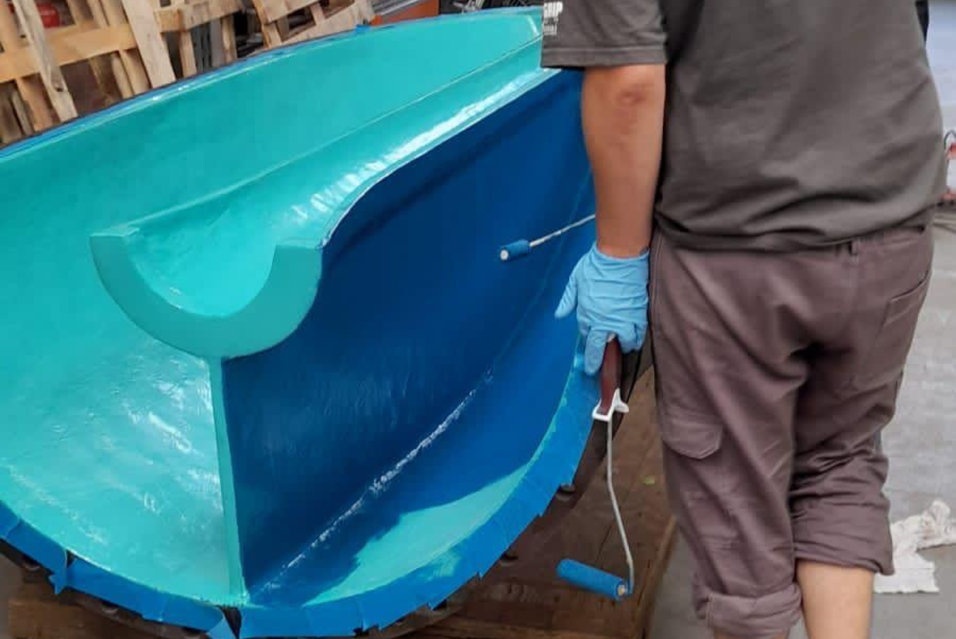 A technician wearing blue protective gloves uses a paint roller to apply a blue topcoat layer onto the inner surface of a large, semi-circular industrial pump housing component already coated in turquoise-green