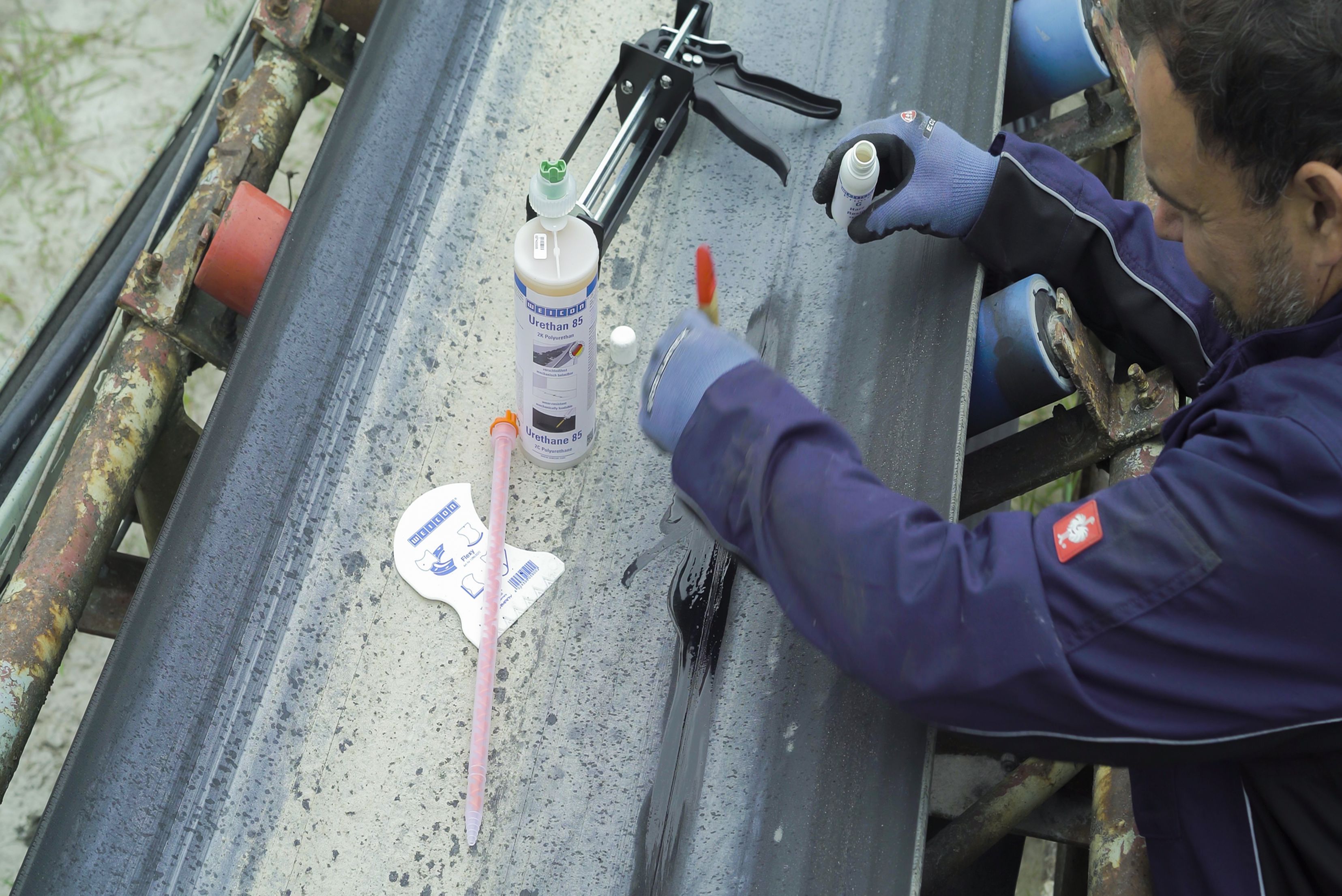 Overhead view of a technician wearing protective gloves applying a black rubber repair resin with a brush to a damage site on an industrial conveyor belt, with a dispenser gun and cartridge resting nearby.