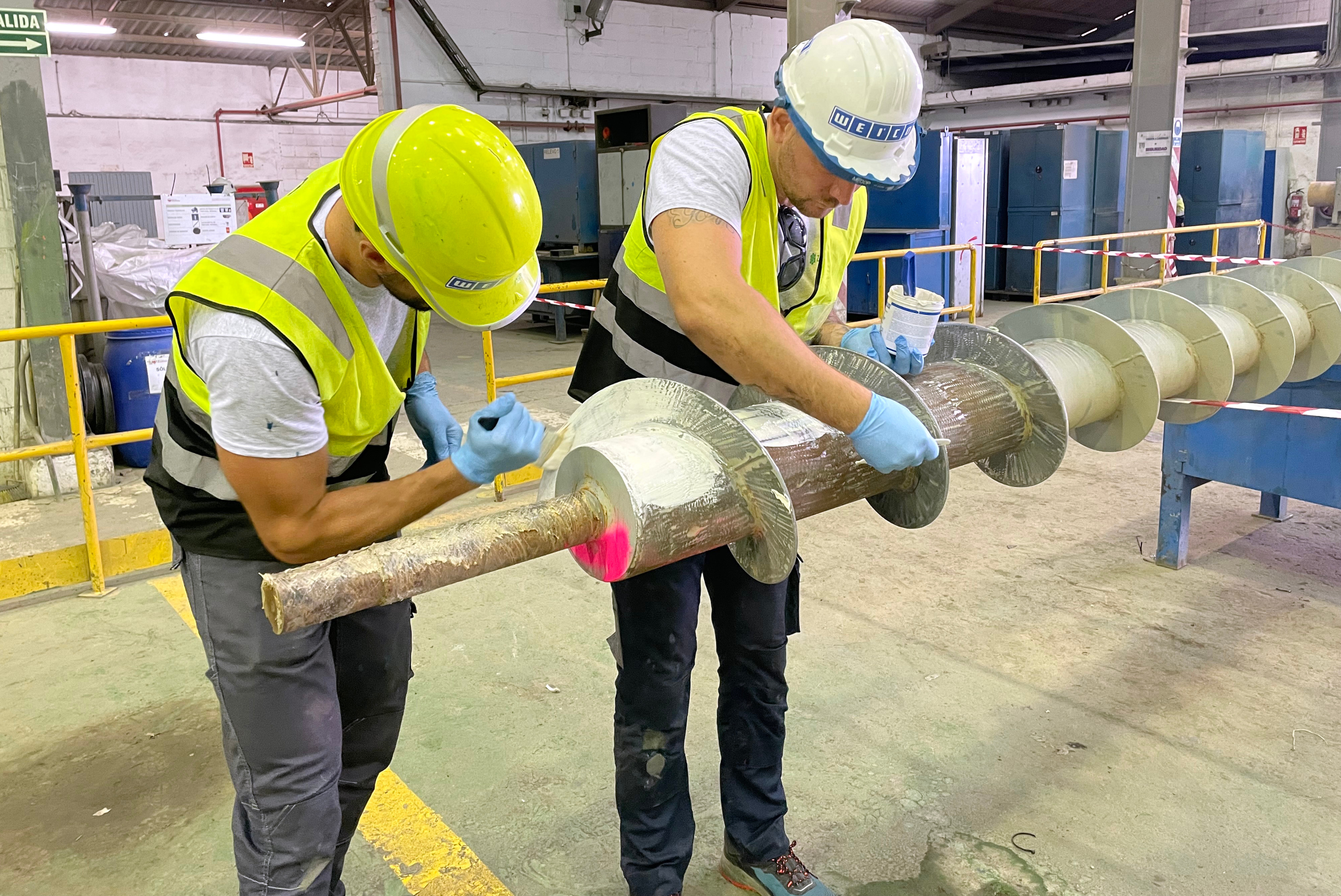 Two technicians wearing hard hats, high-visibility vests, and protective gloves using brushes to manually apply a light-colored protective coating to the flights and shaft of a large industrial screw conveyor in a workshop facility.