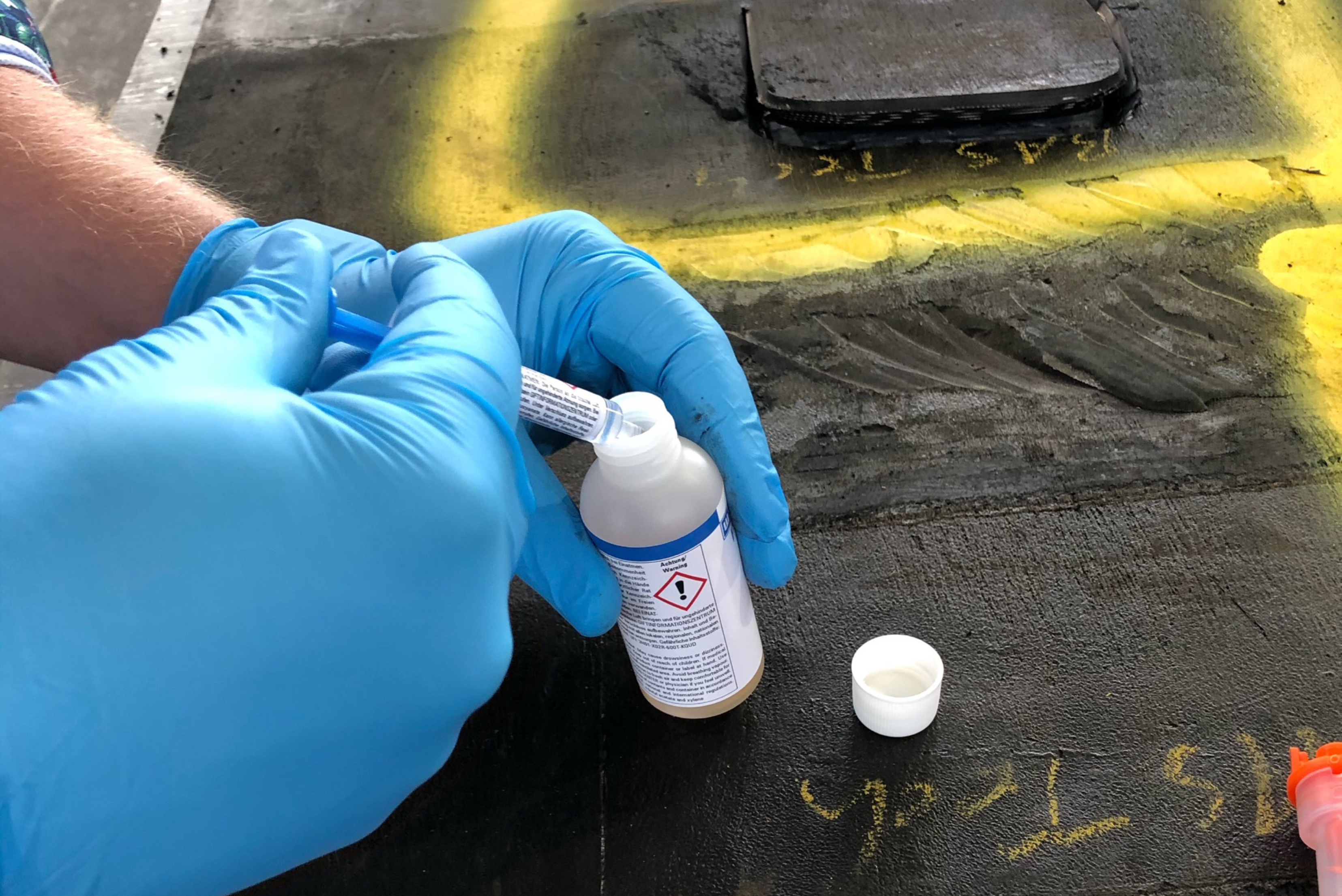Close-up of a person wearing blue protective gloves using a small syringe to dispense liquid into an opened bottle bearing a hazard symbol, resting on a work surface marked with yellow lines.