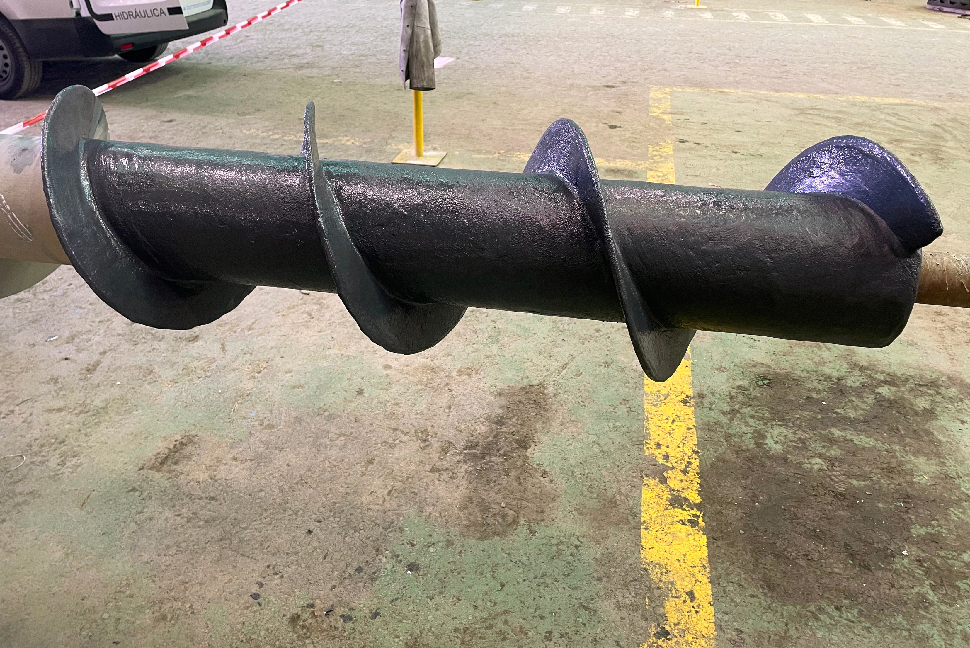 Close-up shot of a large industrial screw conveyor with its central shaft and helical flights completely covered in a smooth, dark protective coating, resting on a workshop floor.