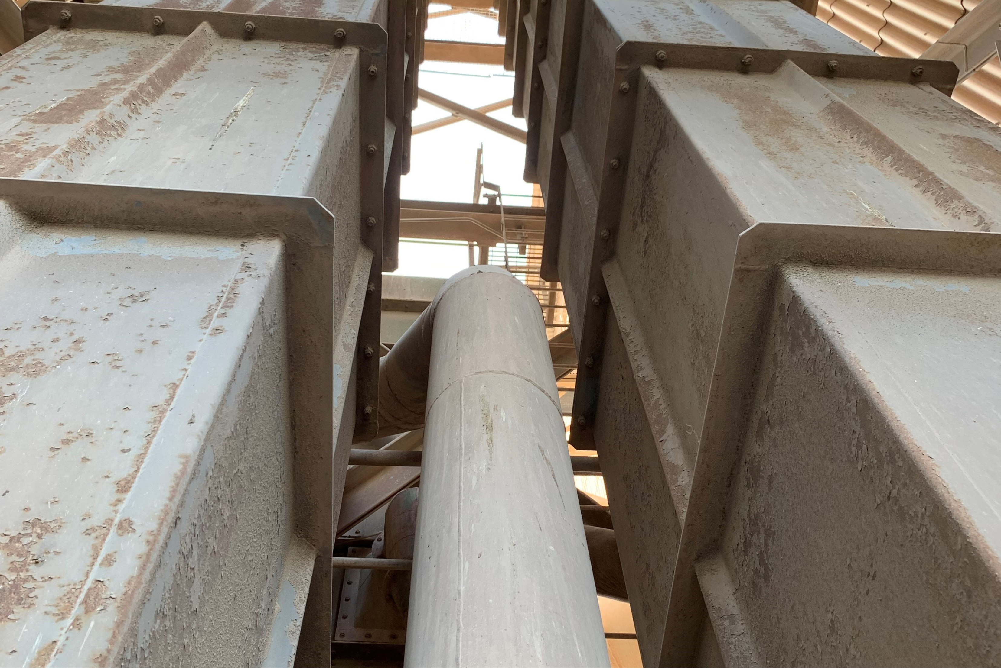 Low-angle view looking up at two vertical, corroded steel bucket elevator shafts with flanged connections and a central process pipe inside an industrial plant.