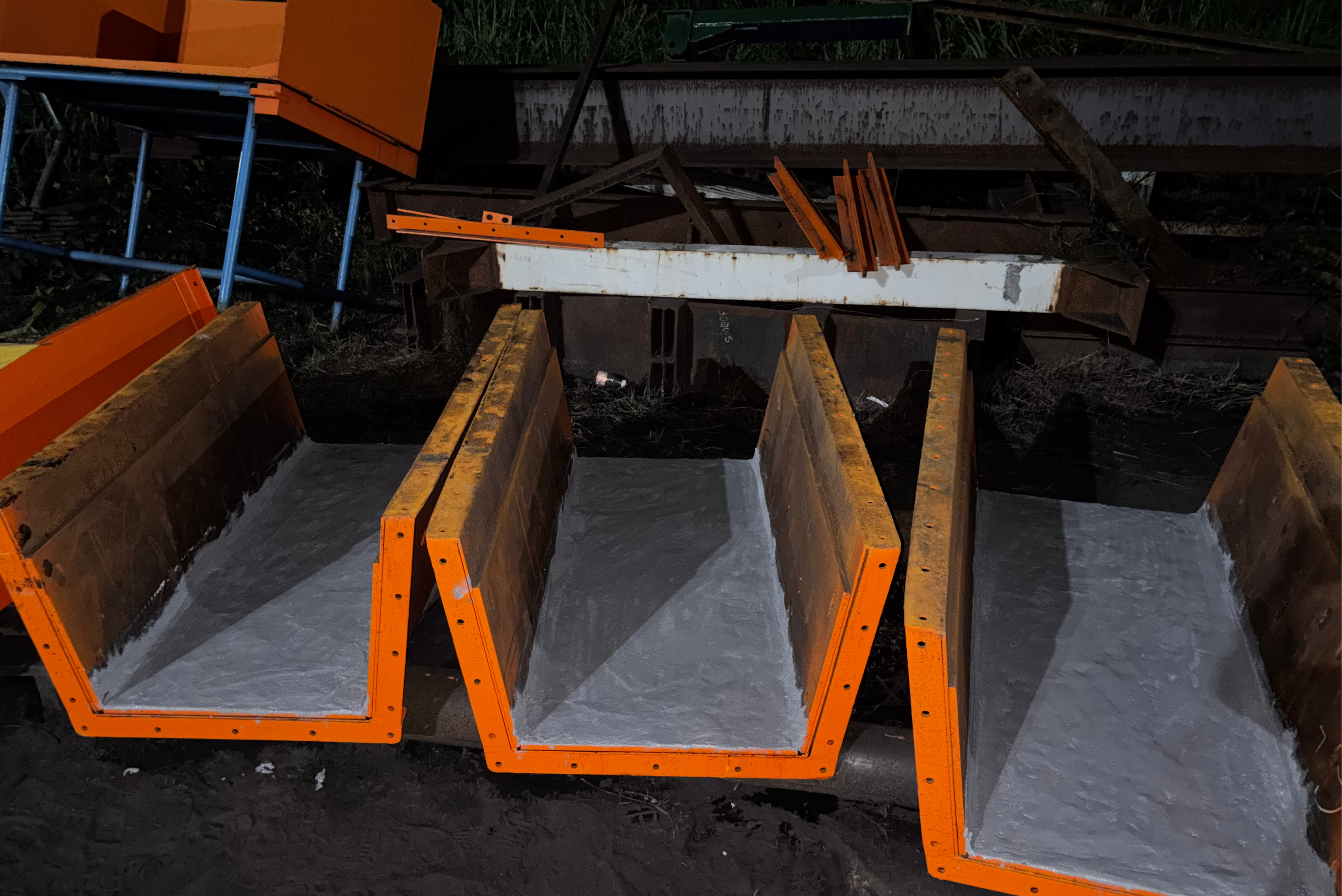 Row of orange-framed heavy steel chute segments lined with a smooth, grey protective coating on their interior surfaces, placed outdoors near industrial steel structures
