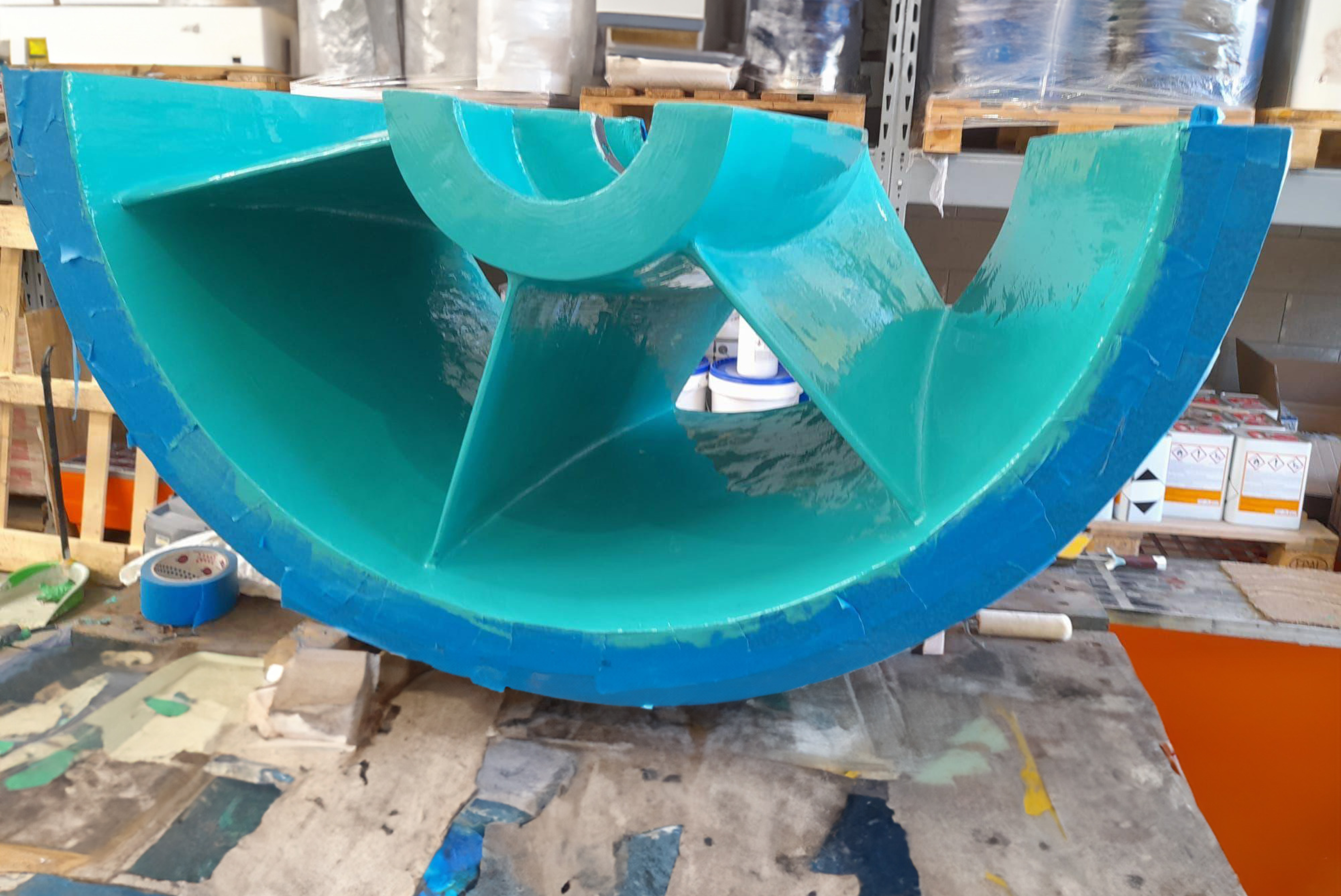 Front view of a semi-circular industrial pump housing component resting on a workbench, featuring a newly applied turquoise-green protective coating on its interior surfaces and blue painter's tape masking along the outer flange edge.