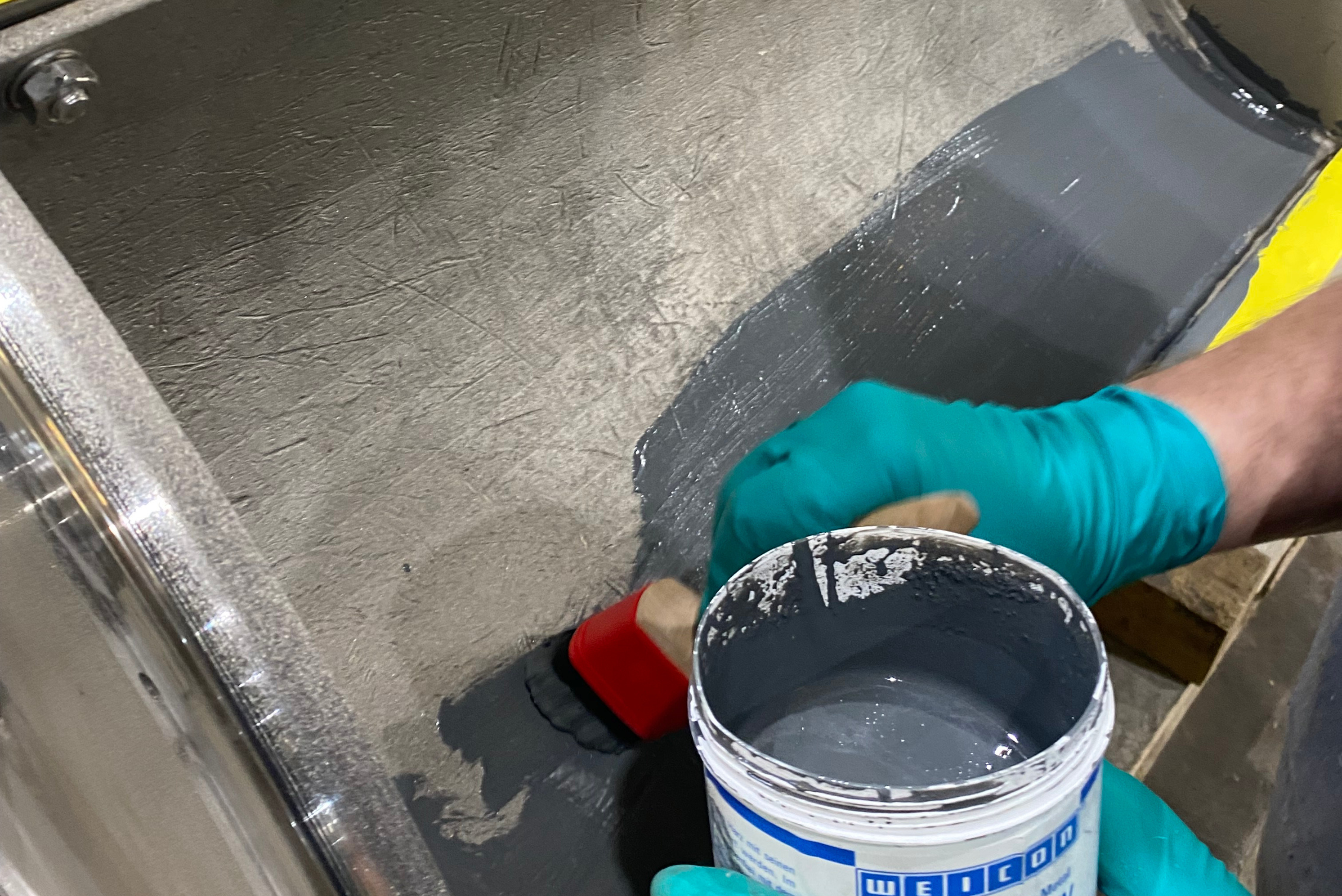 Close-up view of a technician wearing green protective gloves using a paintbrush to apply a grey protective coating from a container onto the internal metallic surface of an industrial component.