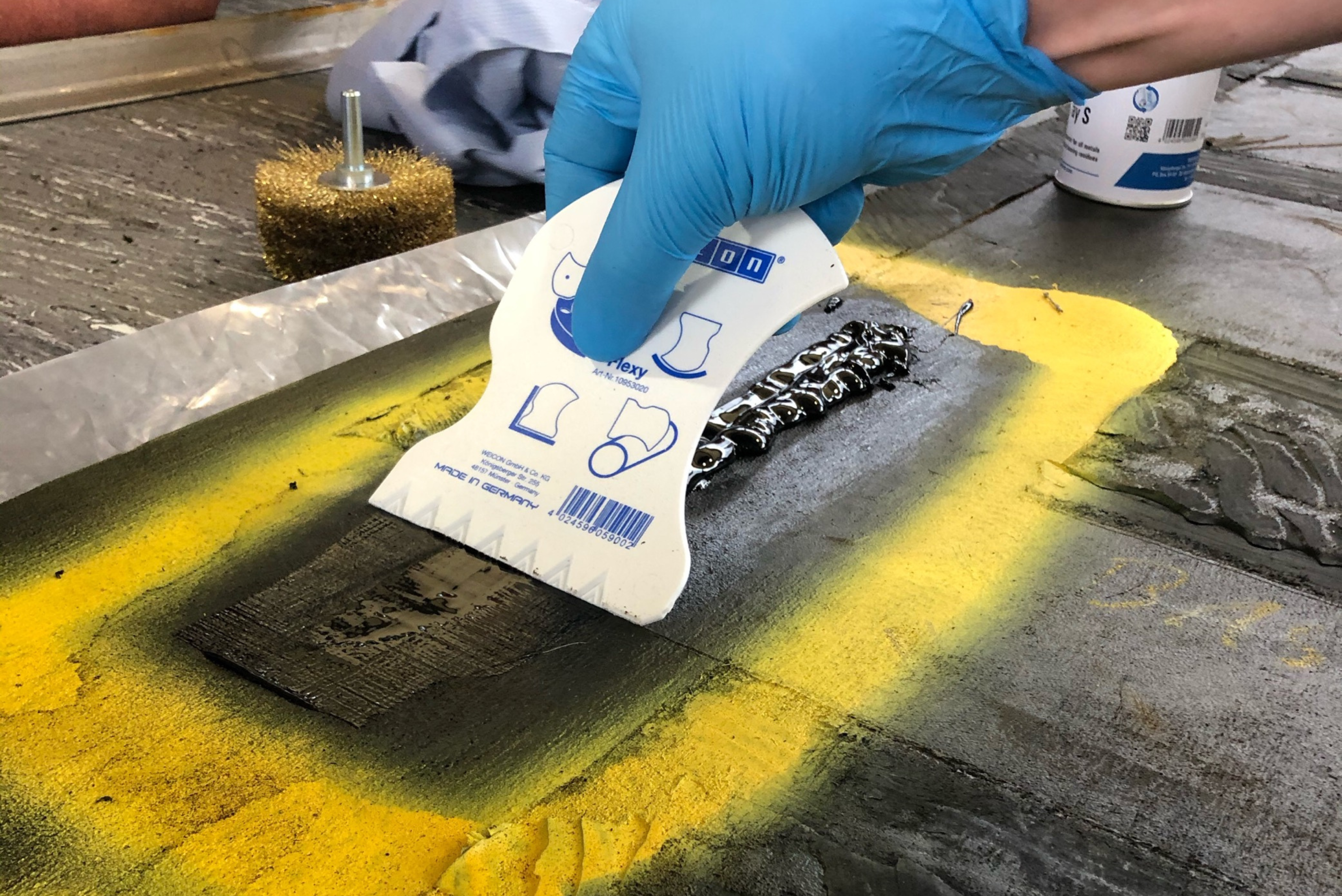 Close-up of a person wearing blue gloves using a plastic spatula to smooth or spread dark sealant across a marked surface.