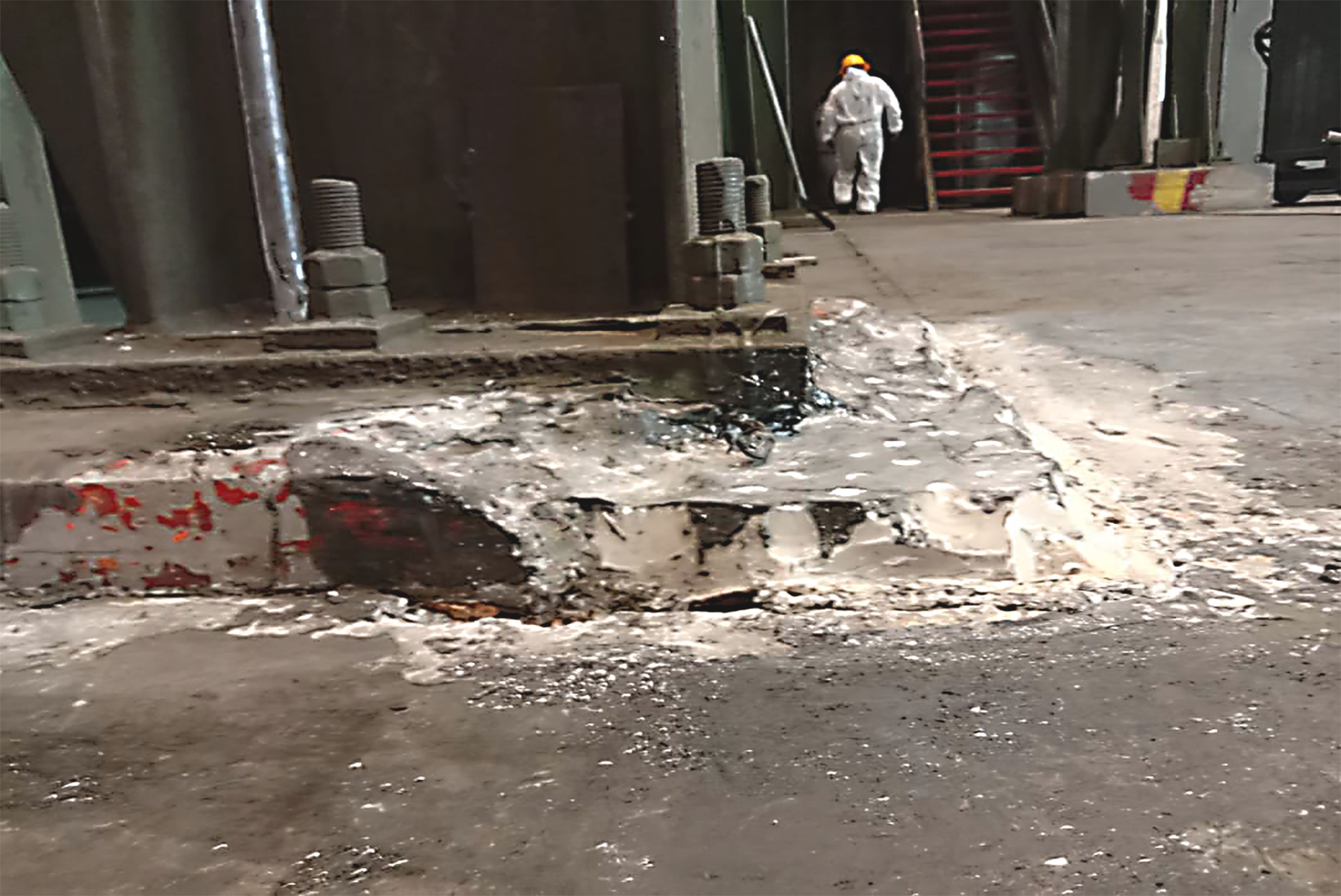 Nahaufnahme der massiven Maschinenbasis und großer Schraubverbindungen auf einem Betonfundament in einer Werkshalle, während im Hintergrund eine in Schutzanzug und Helm gekleidete Person zu sehen ist.
