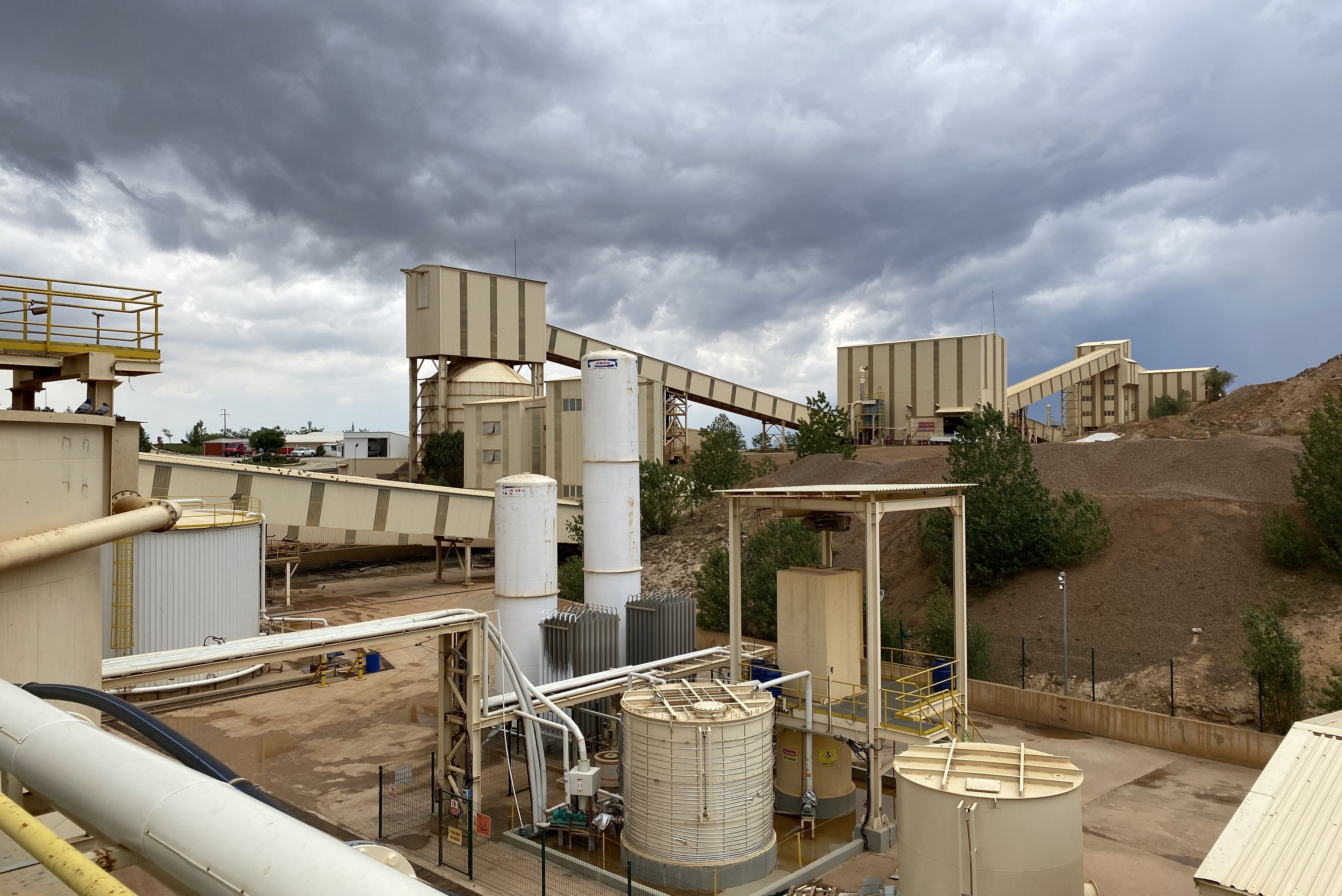 A wide shot of an industrial mineral processing site featuring white storage tanks, vertical silos, connecting conveyor bridges, and processing buildings under a cloudy sky.