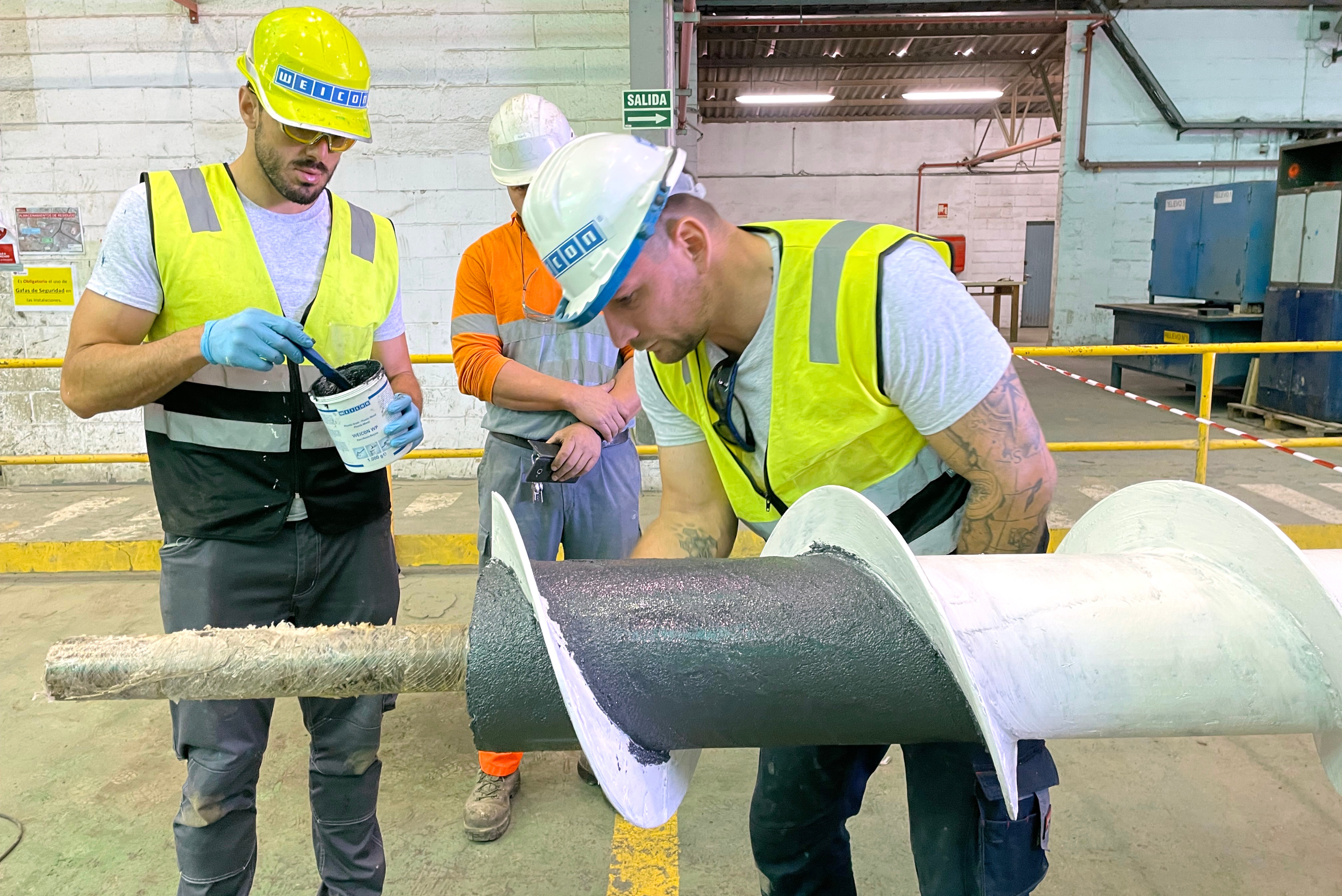 Two technicians wearing protective equipment work on a screw conveyor: while one worker mixes a dark grey coating compound in a small bucket, his colleague carefully applies the material onto the conveyor shaft.